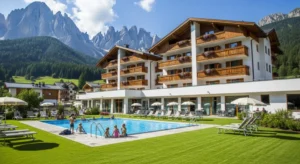 Veduta panoramica di un resort di lusso in montagna con facciata in legno, piscina esterna e prati verdi sullo sfondo delle Dolomiti