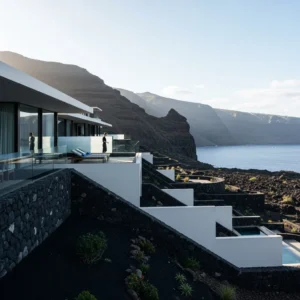 Architettura moderna di un resort con linee pulite, integrato in un paesaggio vulcanico drammatico, con una persona di spalle che ammira la vista.