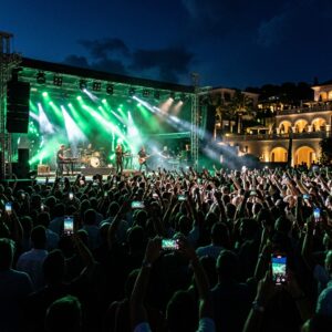 Persone di spalle che assistono a un concerto serale all'aperto con luci verdi e bianche su un palco, durante l'inaugurazione di un resort.