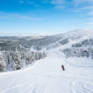 Pista da sci immacolata nel resort di Levi, Finlandia, con cannoni da neve in lontananza e un singolo sciatore visto di spalle che scende la pista tra pini innevati.