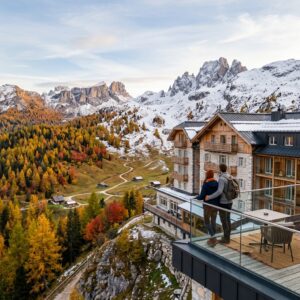 Un lussuoso resort sulle Dolomiti che mostra il passaggio dal foliage autunnale alla prima neve, con una coppia vista di spalle che ammira il panorama dal balcone.